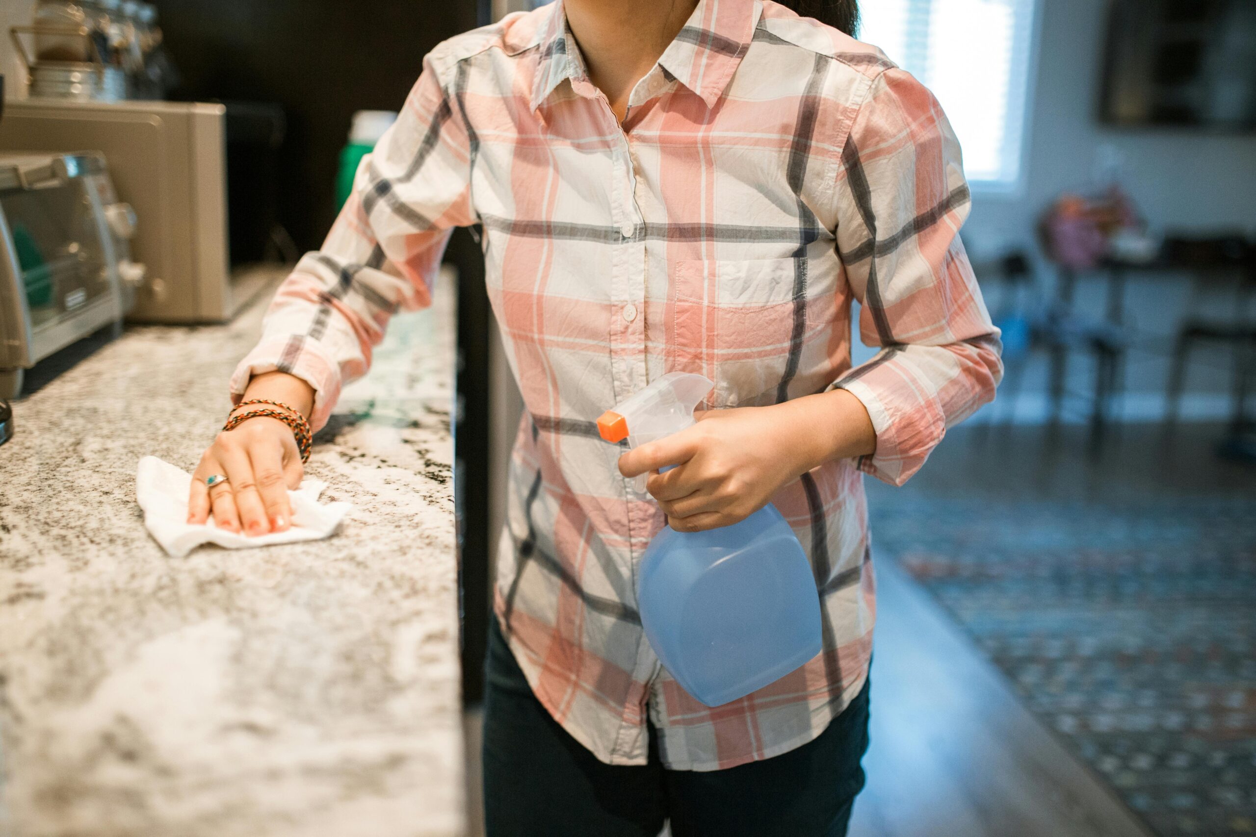 A woman wearing a plaid shirt cleans a kitchen counter using a spray bottle and cloth.